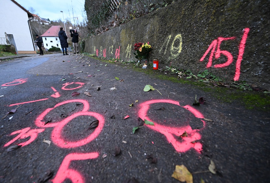 Blumen und Kerzen stehen an Markierungen der Spurensicherung der Polizei an einem Tatort auf einem Weg.