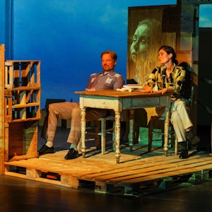 Szene aus „Annette, Ein Heldinnenepos“ im Freien Werkstatt Theater. Annette liegt auf einer Matte am Boden, darüber, auf einer Konstruktion aus Palletts, sitzen ein Mann und eine Frau an einem Tisch.