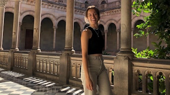 Lena Schwabe auf einem Gang in ihrer Universität in Barcelona. Der Boden ist schwarz und weiß gefliest. Schwabe lächelt in die Kamera. Hinter ihr stehen Säulen und ein Geländer.