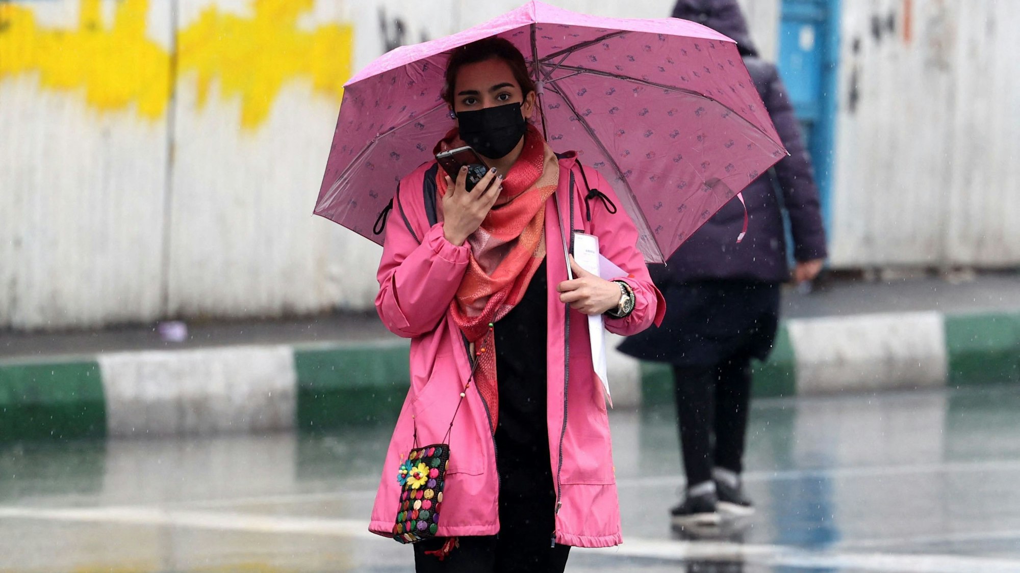 Eine junge iranische Frau läuft ohne Kopftuch an einem Regentag über die Straßen Teherans. Sie trägt einen Mund-Nasen-Schutz und hat einen rosafarbenen Regenschirm dabei.
