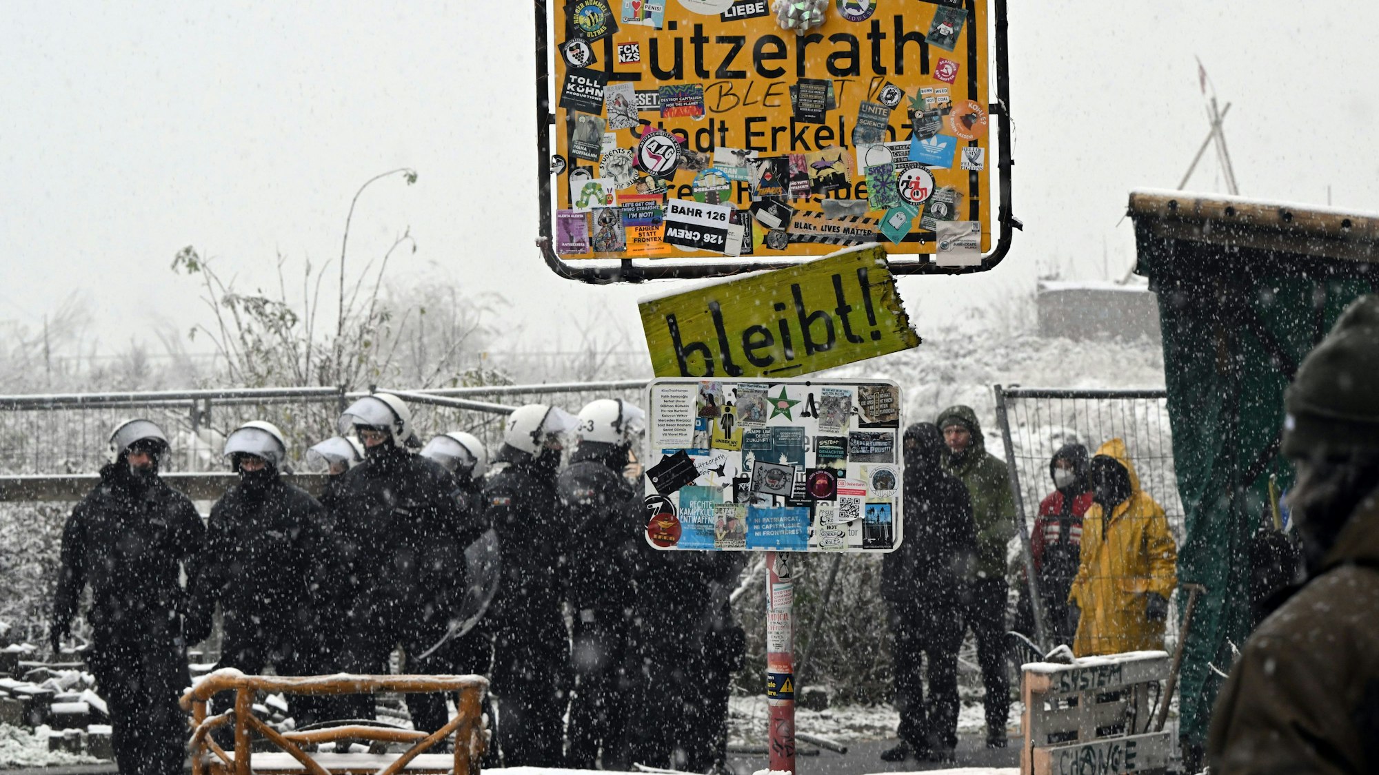 Die Polizei und Aktivisten der Initiative "Lützerath lebt!" stehen vor dem besetzten Gebiet hinter dem Ortsschild, während Schnee fällt.