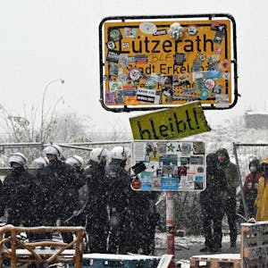 Die Polizei und Aktivisten der Initiative "Lützerath lebt!" stehen vor dem besetzten Gebiet hinter dem Ortsschild, während Schnee fällt.&nbsp;