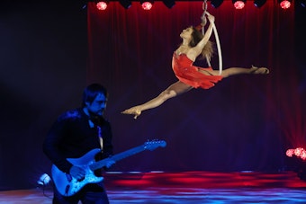 Lady in Red: Eine Artistin im roten Kleid schwebt im Spagat in einem Reifen, während unten am Boden ein Gitarrist ein Solo spielt.