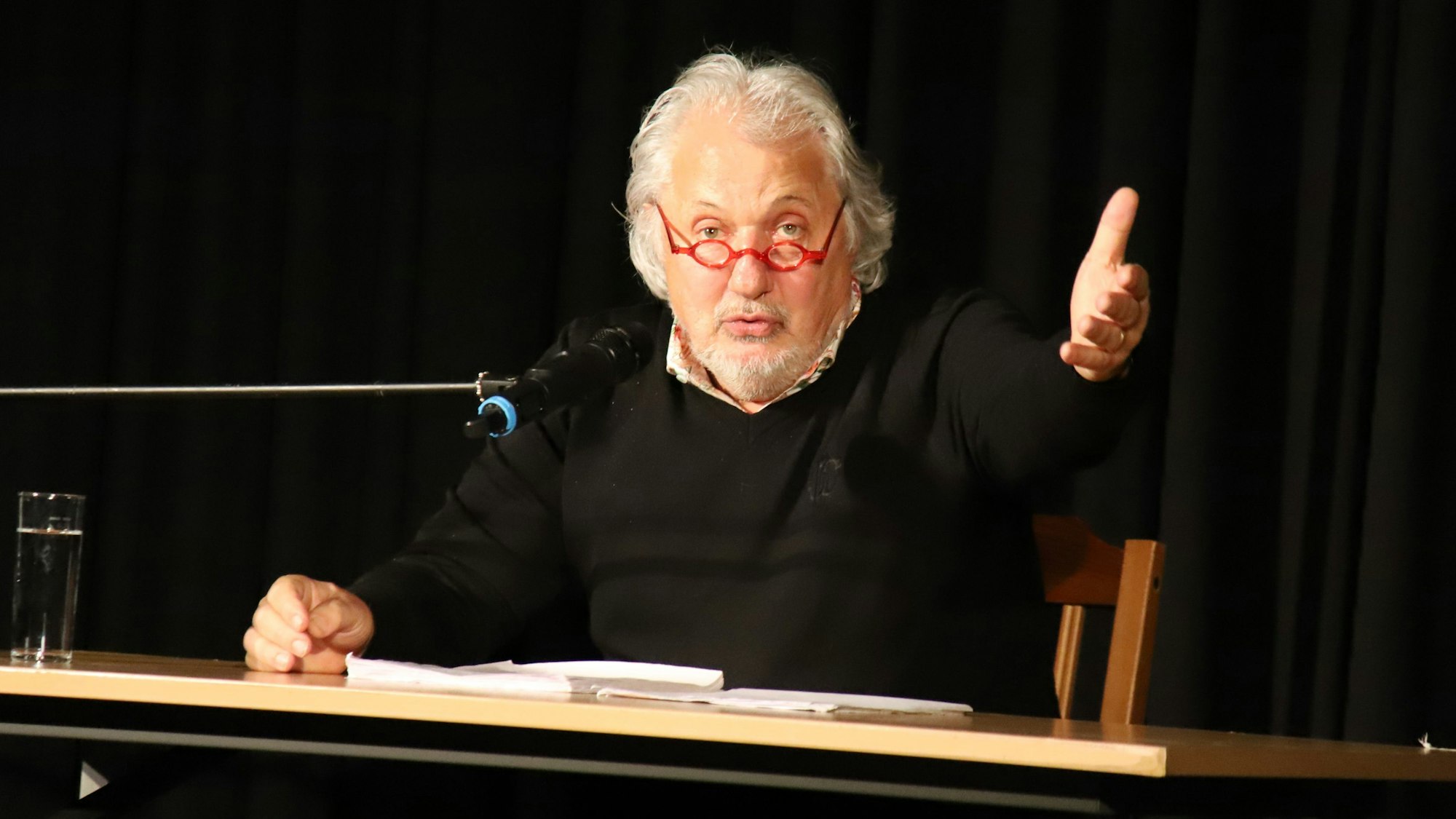 Konrad Beikircher sitzt vor einem Tisch und spricht in ein Mikrofon, die rechte Hand zu einer offenen Geste ausgestreckt. Er trägt eine rote runde Brille.