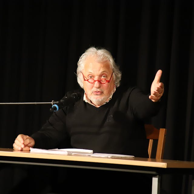 Konrad Beikircher sitzt vor einem Tisch und spricht in ein Mikrofon, die rechte Hand zu einer offenen Geste ausgestreckt. Er trägt eine rote runde Brille.