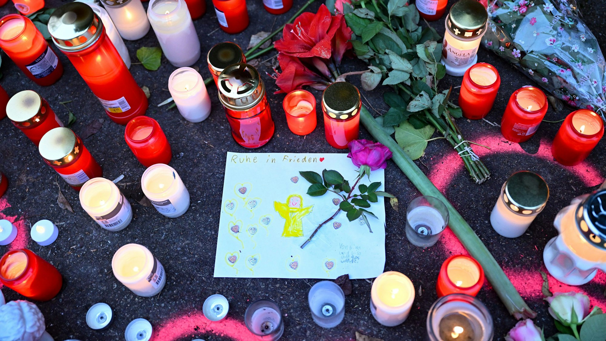 Kerzen und Blumen stehen an einem Tatort, an dem am Tag zuvor zwei Mädchen von einem Mann mit einem Messer angegriffen wurden. In der Mitte ist ein gemaltes Bild von einem Engel, über dem „Ruhe in Frieden“ steht.