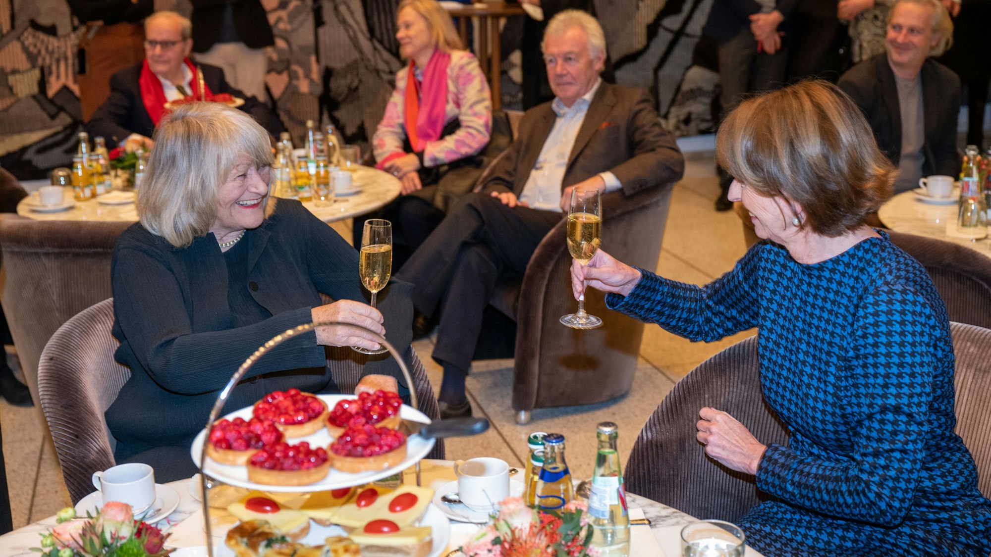 Alice Schwarzer und Henriette Reker am Tisch mit einem Glas Sekt in der Hand.