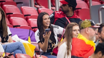 Sky-Moderatorin Laura Winter, hier zu sehen am 1. Dezember 2022 während des WM-Spiels der deutschen Nationalmannschaft gegen Costa Rica in Doha, ist mit DFB-Spieler und Gladbach-Star Jonas Hofmann verheiratet. Winter trägt ein Deutschland-Trikot und hält ein Smartphone in ihrer rechte Hand. Sie lächelt zudem.