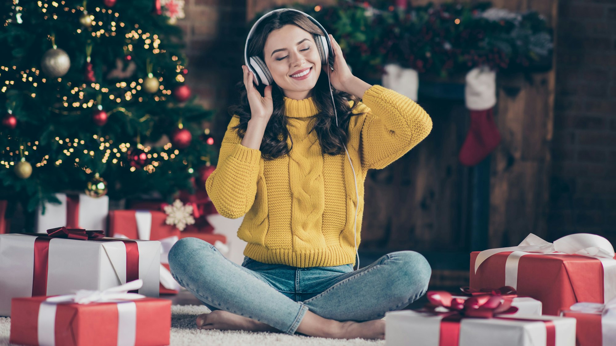 Eine Frau mit Kopfhörern sitzt vor dem Weihnachtsbaum und hat Geschenke vor sich stehen. Sie hört Musik und lächelt mit geschlossenen Augen.