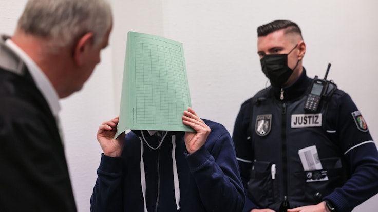 Ein Angeklagter hält sich vor Gericht einen Ordner vor das Gesicht. Neben ihm steht sein Verteidiger und ein Polizist.