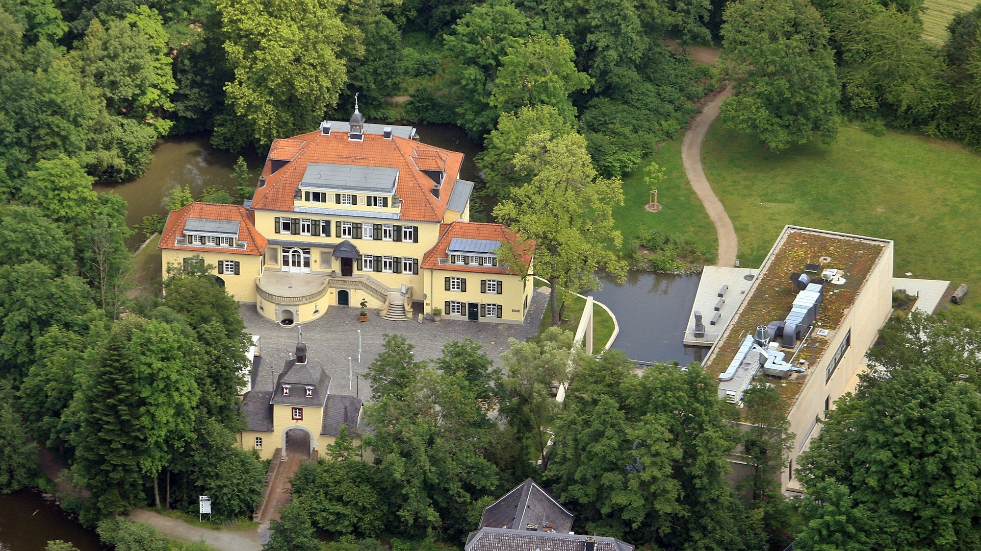 Eine Luftaufnahme von Schloss Eulenbroich inmitten von Waldgebiet und einer Wiesenfläche, die Platz für einen Spielpatz bieten könnte.
