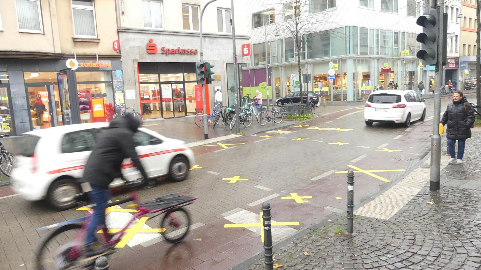 Auf der Venloer Straße sind Markierungen auf Rad- und Fußgängerweg durchgestrichen – es fahren zwei Autos und ein Radfahrer auf der Straße, eine Fußgängerin steht am Straßenrand.