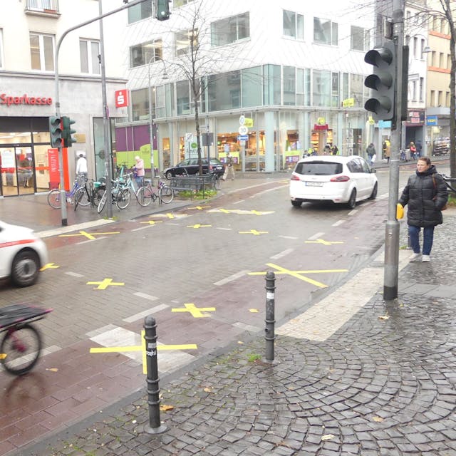 Auf der Venloer Straße sind Markierungen auf Rad- und Fußgängerweg durchgestrichen – es fahren zwei Autos und ein Radfahrer auf der Straße, eine Fußgängerin steht am Straßenrand.
