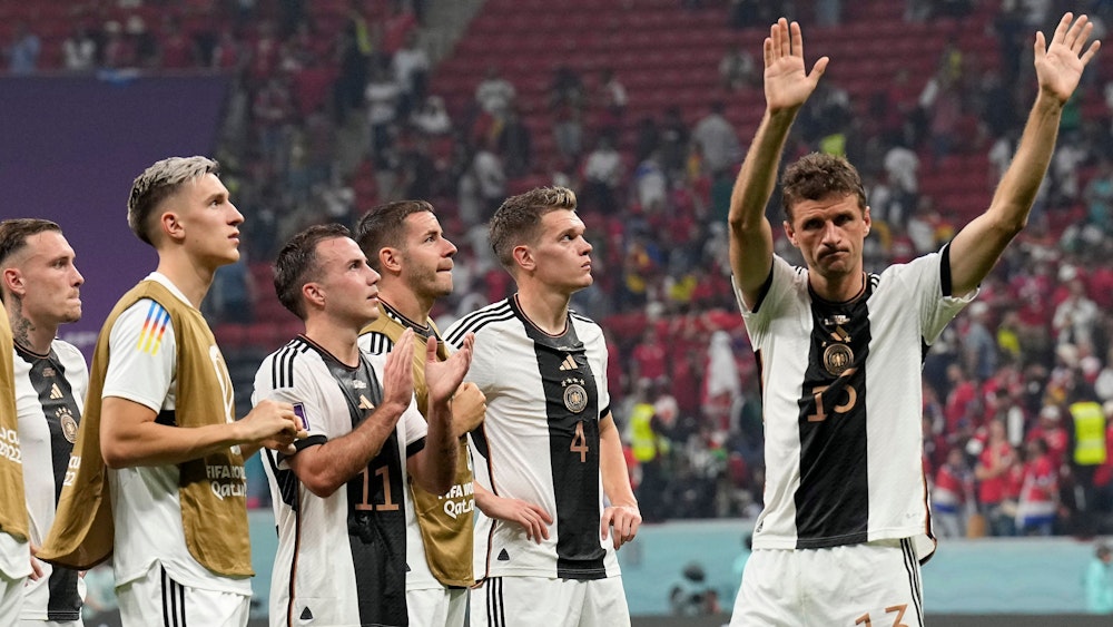 Im Stadion in Katar stehen sechs Spieler der deutschen Nationalmannschaft enttäuscht auf dem Spielfeld vor bereits halbleeren Rängen. Fünf von ihnen schauen nach oben, vermutlich auf die Anzeigentafel, die den Punktestand der Vorrundengruppe zeigt. Rechts winkt Thomas Müller Fans zum Abschied zu.