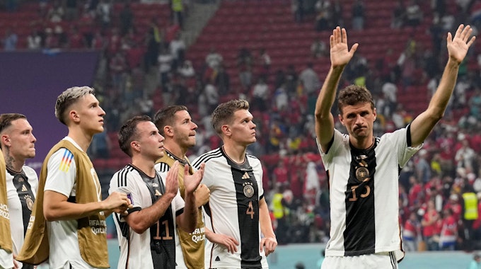 Im Stadion in Katar stehen sechs Spieler der deutschen Nationalmannschaft enttäuscht auf dem Spielfeld vor bereits halbleeren Rängen. Fünf von ihnen schauen nach oben, vermutlich auf die Anzeigentafel, die den Punktestand der Vorrundengruppe zeigt. Rechts winkt Thomas Müller Fans zum Abschied zu.