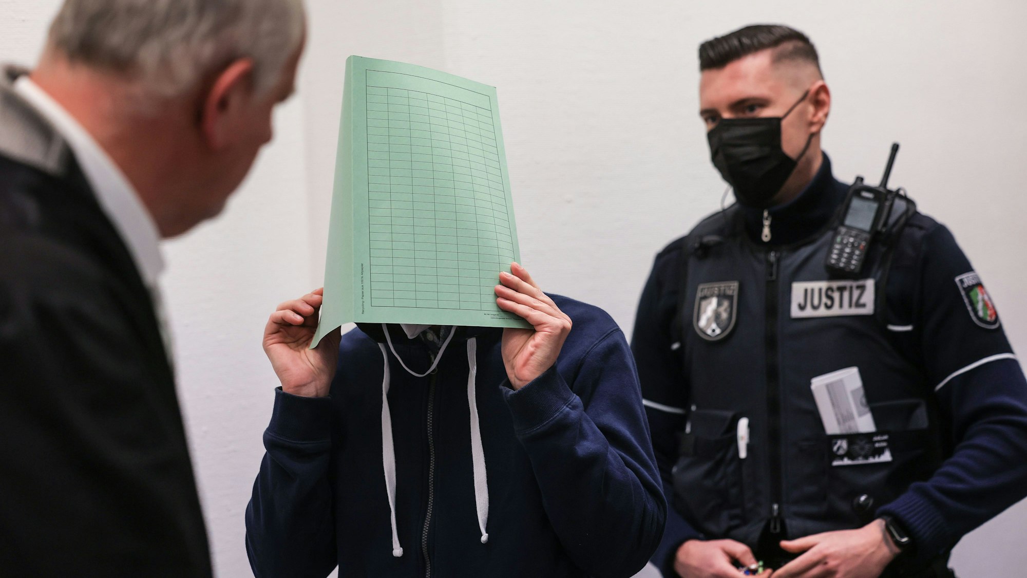 Der Angeklagte (M) im Missbrauchskomplex Wermelskirchen hält sich beim Betreten des Gerichtssaals in Köln neben seinem Rechtsanwalt Christian Lange (l) eine Mappe vor das Gesicht.