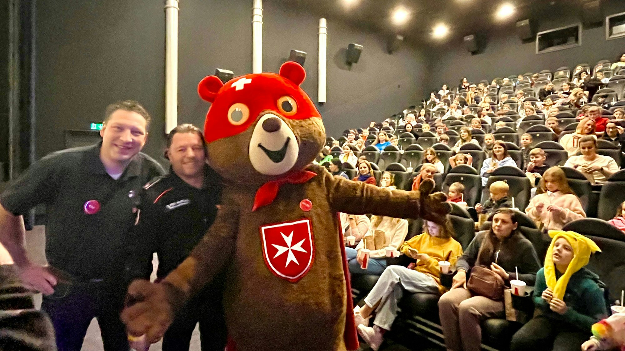 Zwei Männer und ein Mensch im Bärenkostüm vor einem vollen Kinosaal.