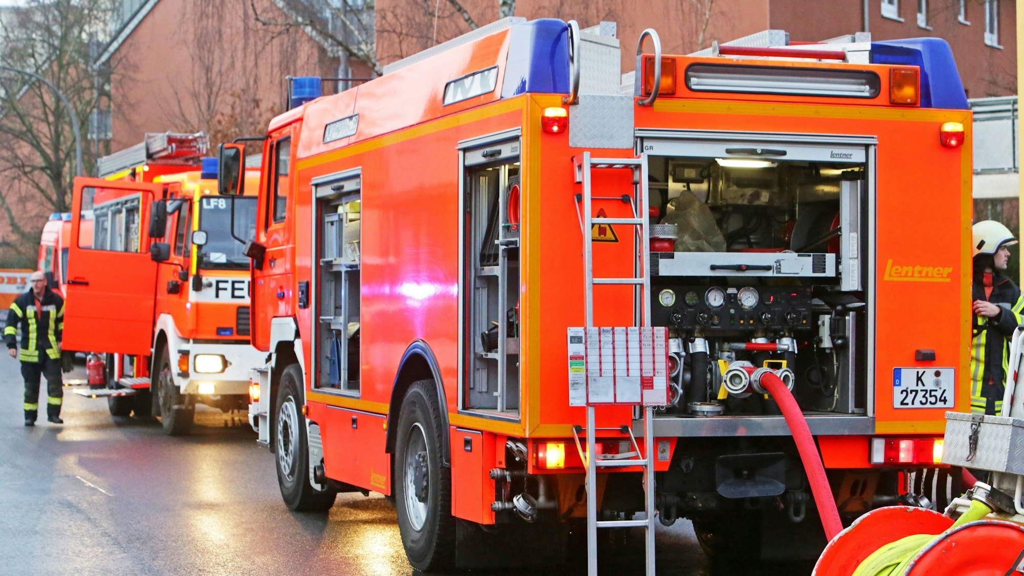 Die Kölner Feuerwehr rückt bei Brandstiftung aus (Symbolbild). Zwei Einsatzfahrzeuge stehen auf der Straße mit zwei Feuerwehrmännern.