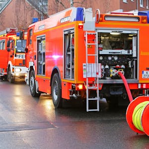 Die Kölner Feuerwehr rückt bei Brandstiftung aus (Symbolbild). Zwei Einsatzfahrzeuge stehen auf der Straße mit zwei Feuerwehrmännern.
