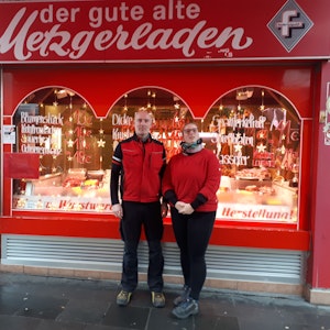 Ein Mann mittleren Alters mit Glatze und eine Frau stehen vor dem in rot gehaltenen Ladenlokal der Metzgerei, die ihnen gehört.
