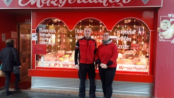 Ein Mann mittleren Alters mit Glatze und eine Frau stehen vor dem in rot gehaltenen Ladenlokal der Metzgerei, die ihnen gehört.