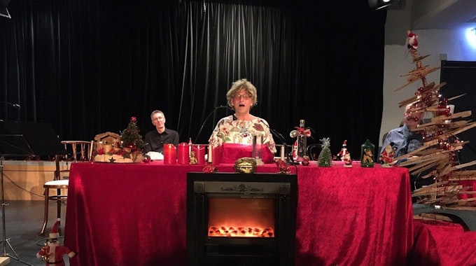 Ein Mann mit Brille sitzt hinter einem altarartigen Tisch mit roter Samtdecke, der weihnachtlich dekoriert ist, neben ihm ist ein Weihnachtsbaum aus Holz zu sehen, im Hintergrund sitzt ein weiterer Mann.