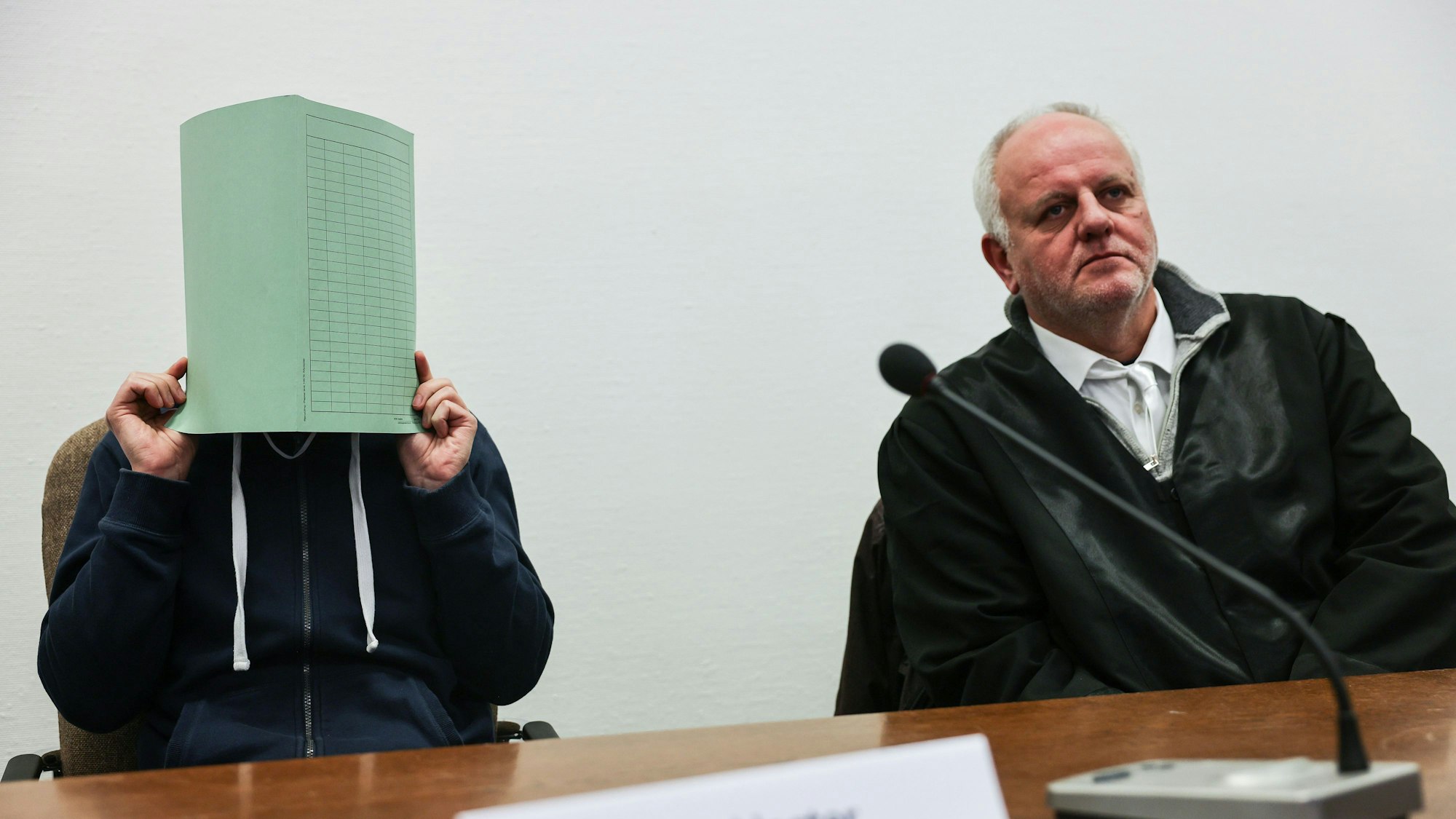 Der Angeklagte (l) im Missbrauchskomplex Wermelskirchen hält sich im Gerichtssaal neben seinem Rechtsanwalt Christian Lange (r) eine Mappe vor das Gesicht.