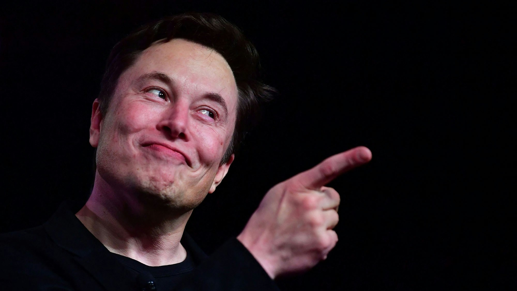 Twitter-Chef Elon Musk steht vor schwarzem Hintergrund, er zeigt mit der rechten Hand.