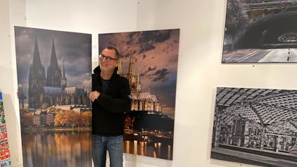 Ein Mann in einer schwarzen Jacke steht rechts rechts vor dem Bild in einer Galerie. Das Gemälde zeigt den Kölner Dom mit seiner Spiegelung im Wasser