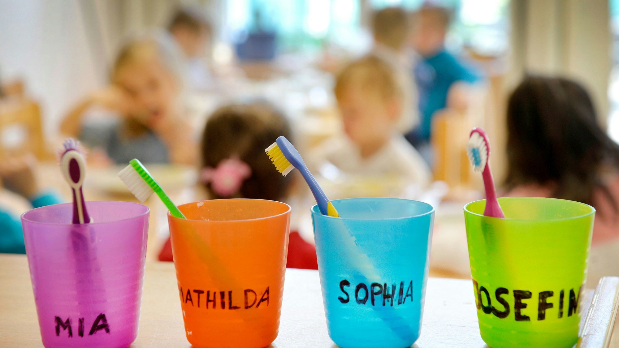 Zahnbürsten und Zahnputzbecher mit den Vornamen der Kinder stehen in einer Kindertagesstätte auf einem Tisch. Im Hintergrund sieht man verschwommen spielende Kinder.
