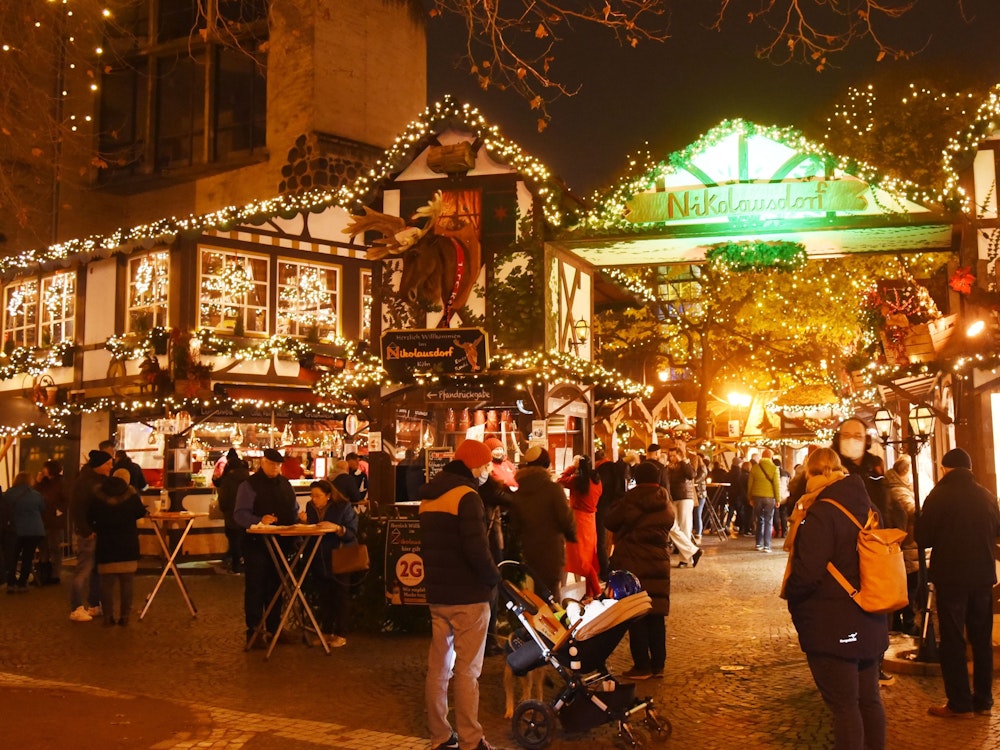 Auf dem Weihnachtsmarkt auf dem Kölner Rudolfplatz stehen viele Menschen und trinken Glühwein oder Punsch. Andere essen oder laufen über den Markt.