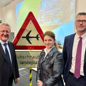 Hans-Peter Zimmermann mit seinen Nachfolgern Anja Mitrenga-Theusinger und André Schumann vor einem Schild: Hubschrauber-Landeplatz
