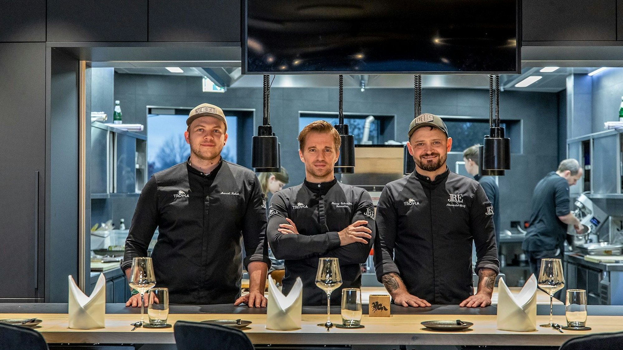 drei Männer in schwarzer Kochkleidung stehen vor einem gedecketen Tisch und blicken in die Kamera