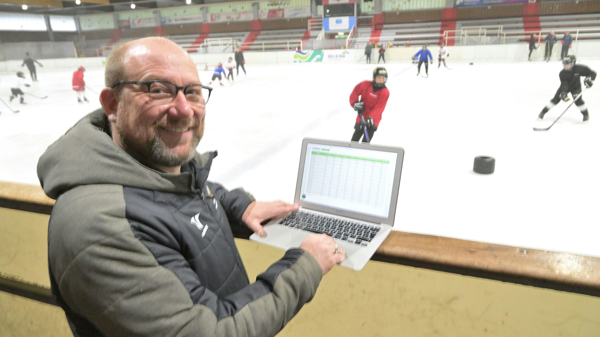 Peter Schüller, Vorstandsmitglied des Eissportvereins Bergisch Gladbach, steht mit Laptop am Rand der Eisbahn, während darauf Jugendliche Eishockey spielen.