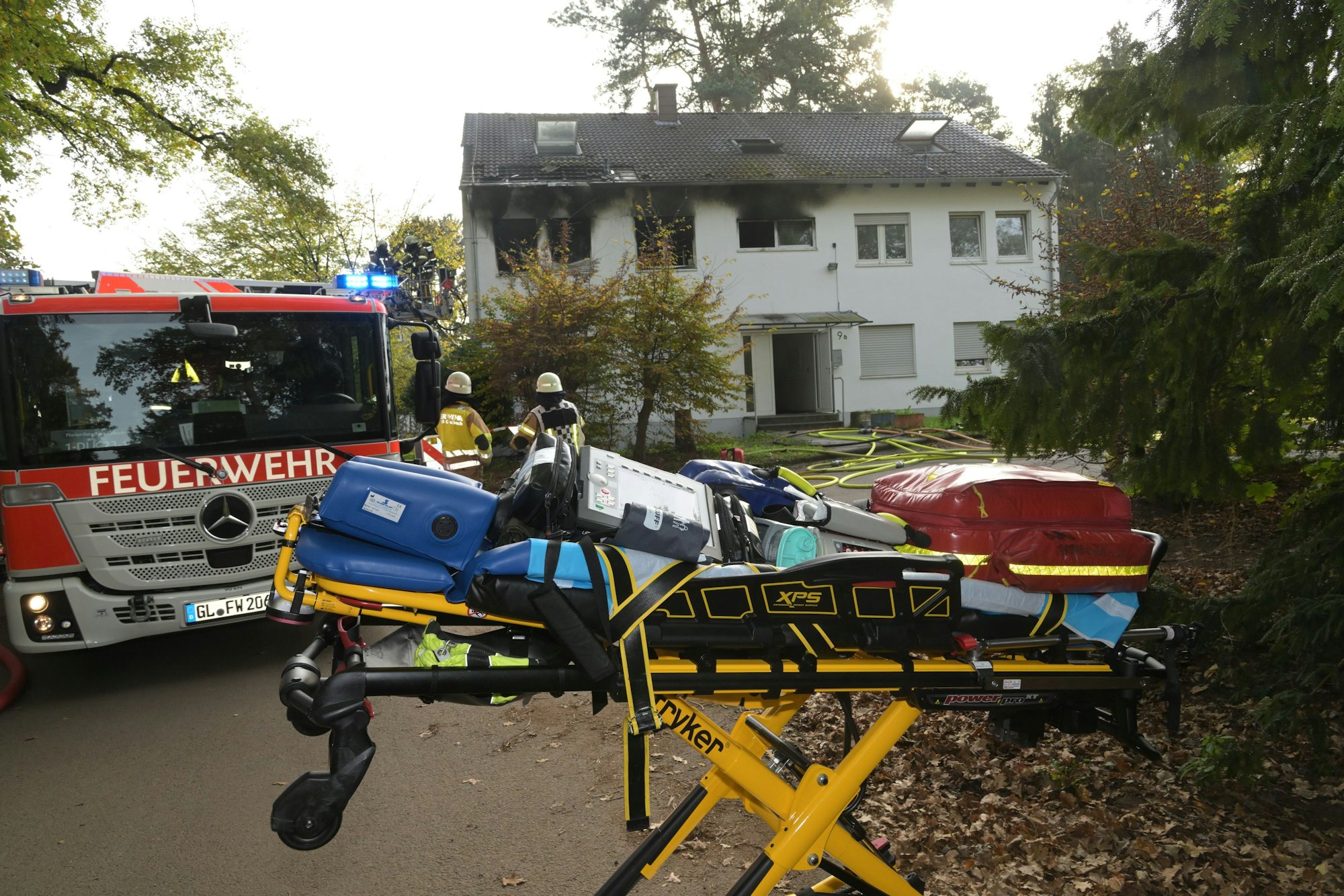 Rauch strmöt aus Fenster der Notunterkunft in Bergisch Gladbach, davor steht ein Feuerwehr und eine Bare mit Gegenständen darauf.