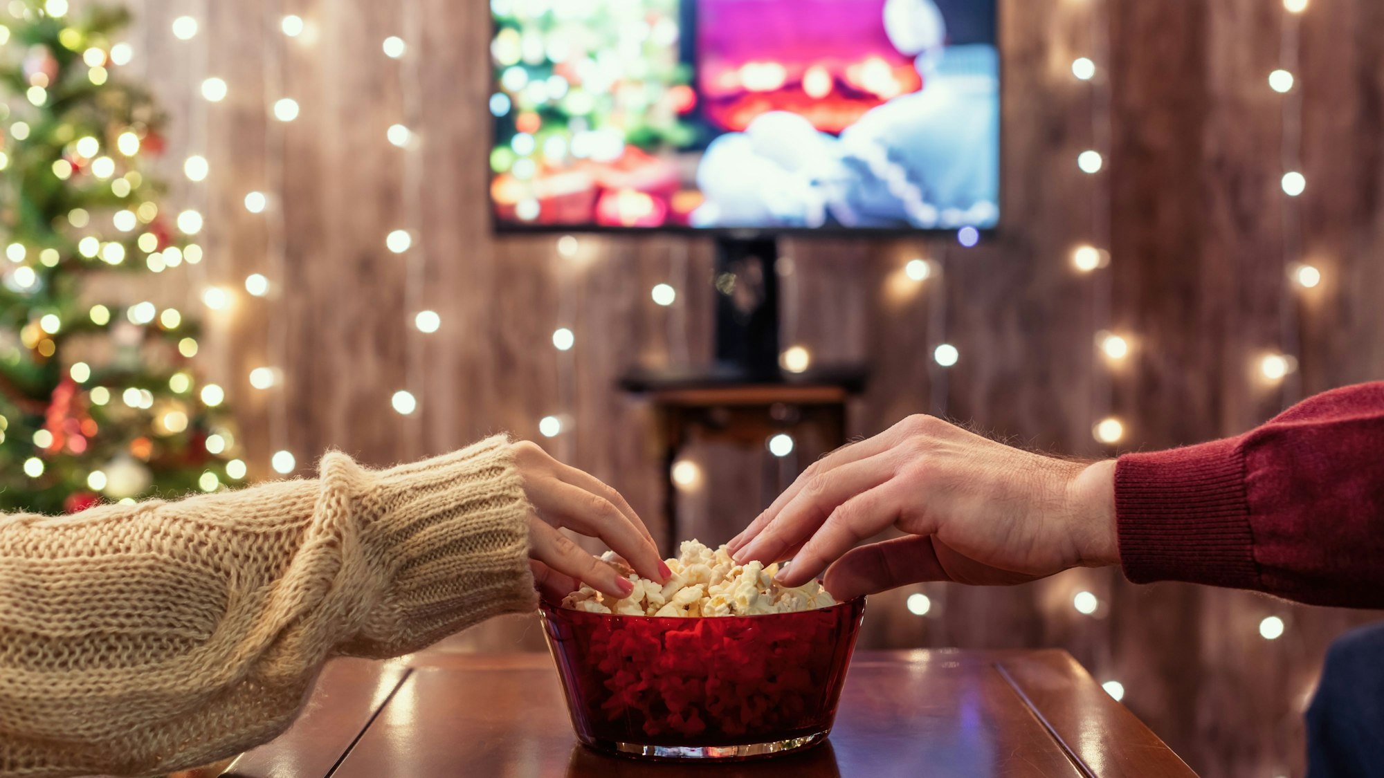 Zwei Hände greifen in eine Popcorn-Schüssel. Im Hintergrund sind verschwommen ein Fernsehen, ein Weihnachtsbaum und Lichterketten.