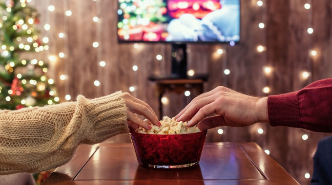 Zwei Hände greifen in eine Popcorn-Schüssel. Im Hintergrund sind verschwommen ein Fernsehen, ein Weihnachtsbaum und Lichterketten.