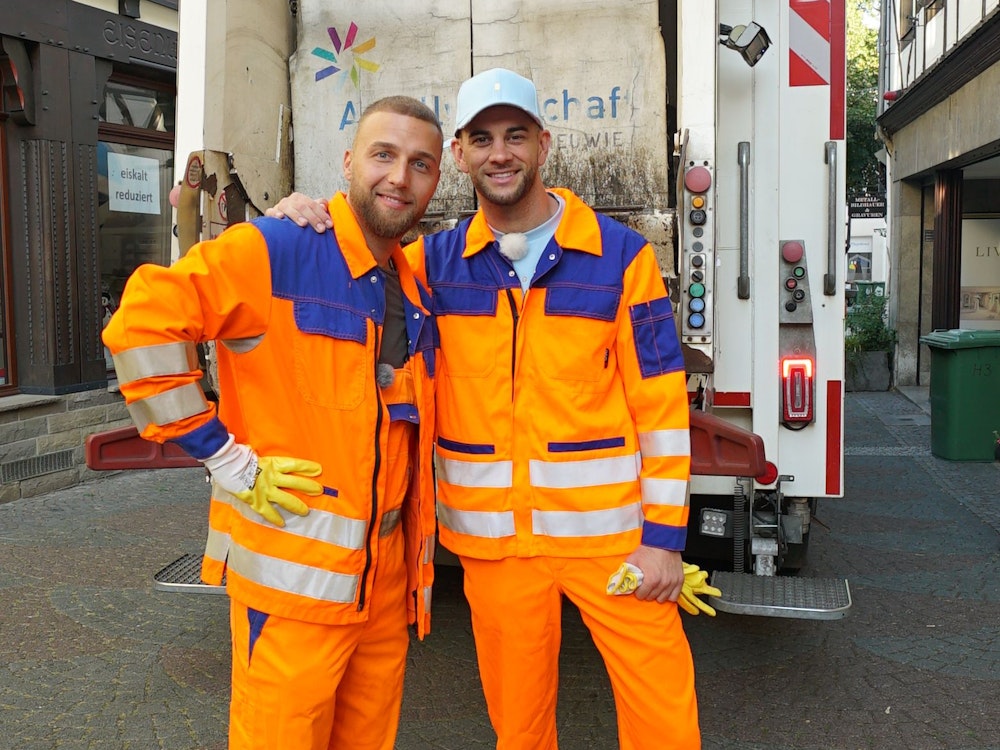 Filip Pavlovic (l.) und Serkan Yavuz tragen ergangene Arbeitskleidung. Sie stehen vor einem Müllwagen.