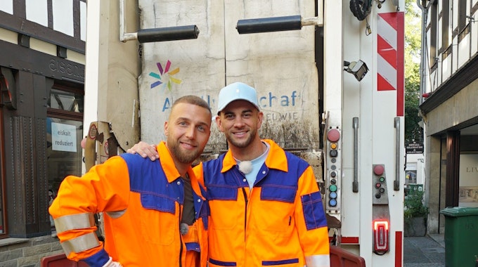 Filip Pavlovic (l.) und Serkan Yavuz tragen ergangene Arbeitskleidung. Sie stehen vor einem Müllwagen.