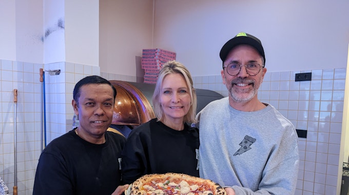Pizzabäcker Monir (l.) posiert am 8. Dezember 2022 mit Chefin Verena Malta und Chef Ingo Noack. Sie halten einen Teller, auf dem sich eine vegane Pizza befindet.