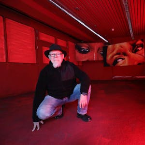 Ein Mann mit Hut kniet in einem komplett roten Raum, im Hintergrund hängen zwei großformatige Fotos, die verängstige Frauenaugen zeigen.