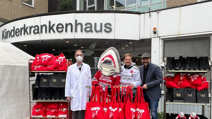 Professor Michael Weiß, Sharky, KEC-Kapitän Moritz Müller sowie Haie-Geschäftsführer Philipp Walter mit den Geschenken vor der Kinderklinik.