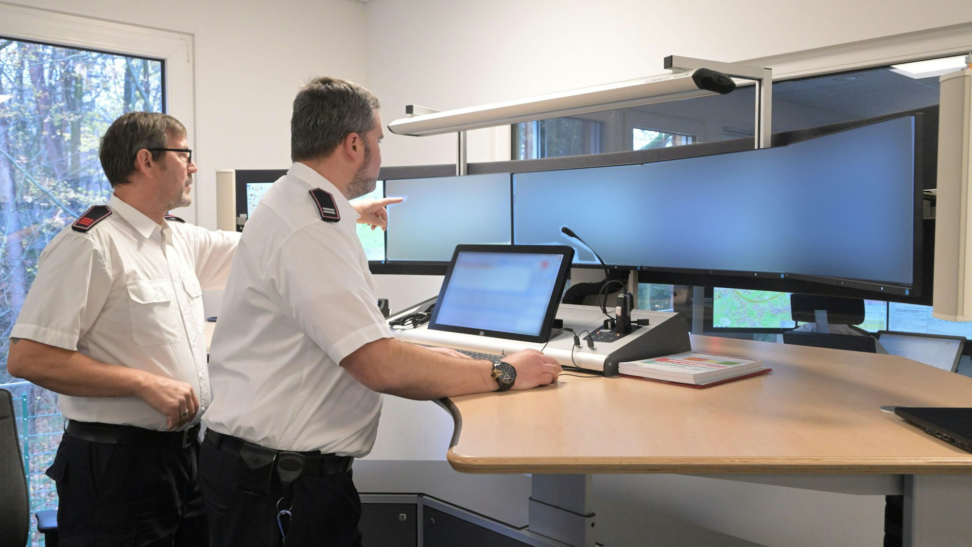 Zwei Mitarbeiter der Kreisleitstelle in weißem Hemd und Uniformhose stehen vor mehreren Bildschirmen an einem Stehpult in der Kreisleitstelle.