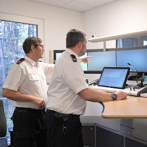 Zwei Mitarbeiter der Kreisleitstelle in weißem Hemd und Uniformhose stehen vor mehreren Bildschirmen an einem Stehpult in der Kreisleitstelle.