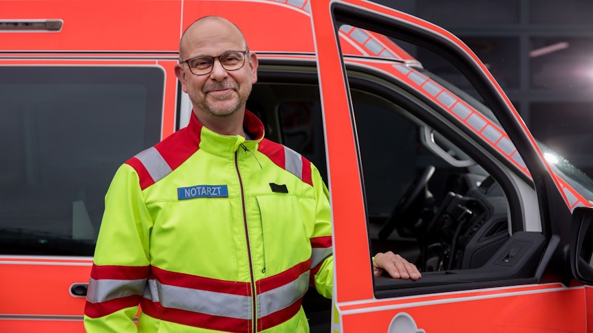 Notarzt Marco Strohm steht in Berufskleidung vor einem Einsatzfahrzeug der Feuerwehr Köln.