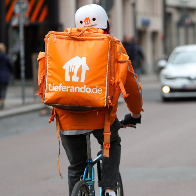Ein Fahrradbote vom Lieferdienst „Lieferando“ fährt durch die Innenstadt. Auf dem Rücken trägt er einen orangefarbenen Rucksack.