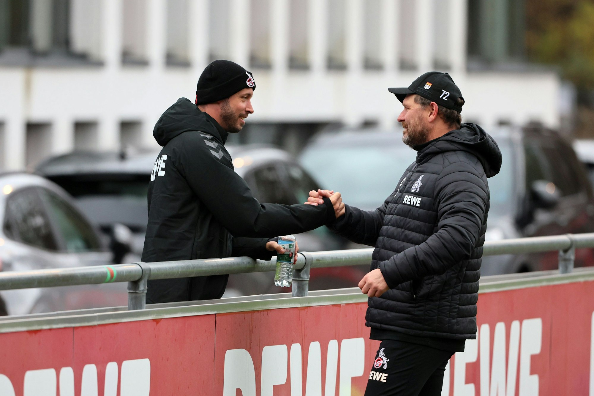 Kölns verletzter Offensivspieler Mark Uth (l.), der eine schwarze Mütze trägt, begrüßt FC-Coach Steffen Baumgart am Rande der Trainingseinheit.