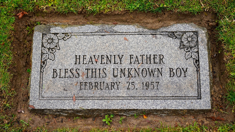 Das Foto zeigt einen grauen Grabstein mit Inschrift und Blumenornamenten.