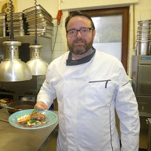Johannes Richter steht in der Restaurant-Küche und posiert mit einem angerichteten Teller für die Kamera.