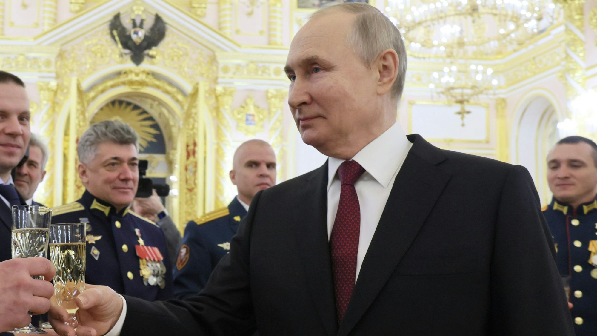 Der russische Staatspräsident Wladimir Putin beglückwünscht mehrere Personen während einer Zeremonie am Vorabend des Tages der Helden des Vaterlandes im Kreml. Er trägt einen Anzug und hat ein Champagner-Glas in der Hand.